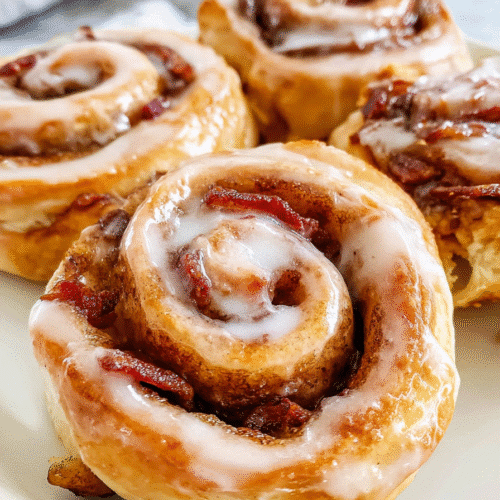 bacon cinnamon rolls topped with melted icing and crispy bacon bits on a white plate